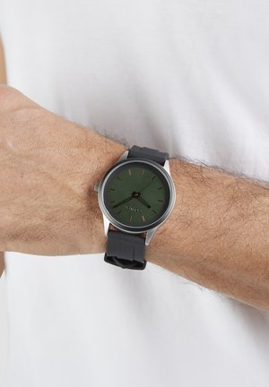 Round watch with a dark green dial, brass hour markers, and a stainless steel case. It has a black rubber strap.