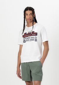 Fehér, rövid ujjú póló piros és sötétkék grafikai nyomattal, amelyen a "Jack & Jones Superior Quality" szöveg szerepel, zöld rövidnadrággal viselve.