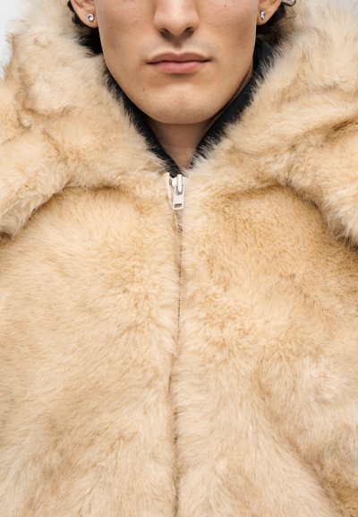 Μπεζ παλτό από γούνα με παχύ, υφή υλικό· διαθέτει φερμουάρ και καπό με σχέδιο για επιπλέον ζεστασιά.