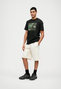 T-shirt en coton noir avec un graphique de logo Lacoste coloré, associé à un short crème et des baskets de sport noires, se tenant devant un fond uni.
