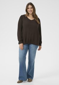 Smilende kvinde med langt hår iført en løs mørkebrun bluse, lyseblå jeans og brune ballerinaer, der står foran en lys baggrund.