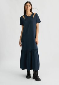 Abito maxi blu navy con maniche corte, caratterizzato da aperture sulle spalle e dettagli in metallo. Fondo svasato con cuciture arricciate. Indossato con stivali neri.
