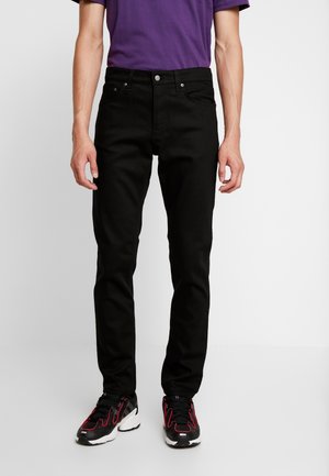 Person iført slim-fit sorte jeans, lilla skjorte og sorte sneakers med røde detaljer, stående mod en ensfarvet lys baggrund.