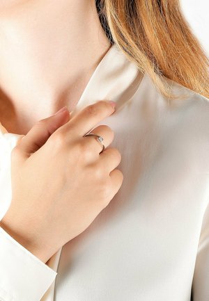 Hand mit Silberring hält den Kragen einer cremefarbenen Bluse, mit hellbraunem Haar, das auf der Schulter sichtbar ist.