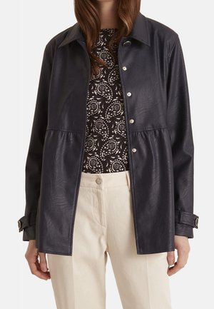 Femme portant une veste en cuir noire boutonnée, un haut à motifs noir et blanc, et un pantalon crème taille haute, debout les bras détendus.