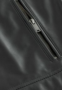 Detail einer schwarzen Lederjacke mit einem Metallreißverschluss, strukturierten Oberflächen und genähten Akzenten rund um den Reißverschlussbereich.