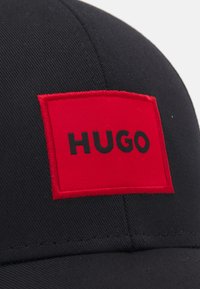 Must värvitud basebollimüts, millel on kangast tempel, kus on julge punane taust ja tõstetud must tekst "HUGO." Sile tekstuur, struktuurne kuju.