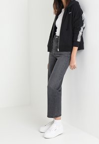 Sudadera negra con cremallera y rayas blancas en las mangas, combinada con vaqueros grises de cintura alta y zapatillas blancas, con un ajuste casual.