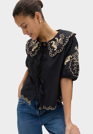 Vrouw draagt een zwarte blouse met beige bloemrijke borduursels, geschulpte randen en opgeblazen korte mouwen, gecombineerd met een blauwe spijkerbroek.