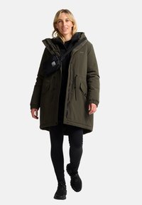 Parka vert olive avec capuche, fermeture éclair et boutons, et poches avant. Associée à des leggings noirs et des baskets. Sac bandoulière noir.