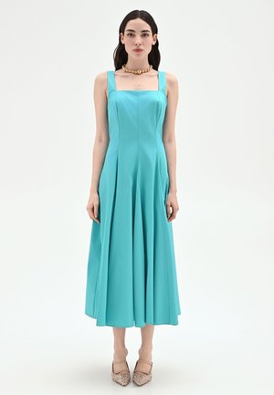 Donna con capelli scuri che indossa un vestito midi turchese senza maniche e tacchi beige con motivo, in piedi su uno sfondo bianco semplice.