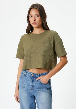 T-shirt corta color verde oliva in tessuto texturizzato con maniche corte e tasca frontale, abbinata a jeans a vita alta di color blu chiaro.