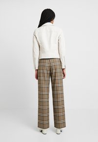 Bílá kožená bunda s přiléhavým střihem, doplněná hnědými vysokým pasem kalhotami v kostkovaném vzoru; model stojí s rukama podél těla.