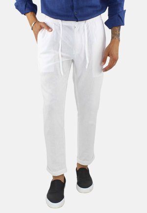 Uomo che indossa pantaloni di lino bianchi con coulisse abbinati a una camicia blu a maniche lunghe e scarpe slip-on nere su sfondo bianco.