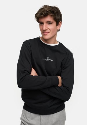 Sweatshirt noir en tissu doux, avec un col rond, des poignets côtelés et un petit logo blanc sur la poitrine. Un pantalon gris complète la tenue.