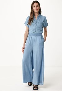 Jasnoniebieski dżinsowy kombinezon z krótkimi podwiniętymi rękawami, zapięciem na guziki z przodu, kieszenią na piersi oraz szerokimi spodniami z elastycznym paskiem w talii.