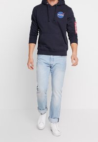 Felpa con cappuccio blu marina con logo NASA, tasca frontale e dettaglio etichetta rossa sulla manica. Jeans azzurri e sneakers bianche completano il look.