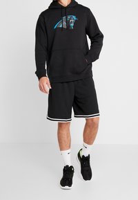 Schwarzer Hoodie mit blauer Panther-Logo, vorne mit Kängurutasche. Kombiniert mit schwarzen Shorts mit weißen Seitenstreifen, weißen Socken und sportlichen schwarzen Schuhen.