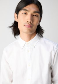 White button-up shirt with a collar, featuring a textured fabric and small embroidered detail on the left chest. Standard fit design.