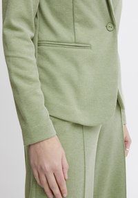 Zbliżenie osoby w jasnoszarym, strukturalnym żakiecie i dopasowanych spodniach, trzymającej jedną rękę wzdłuż ciała na jednolitym tle.