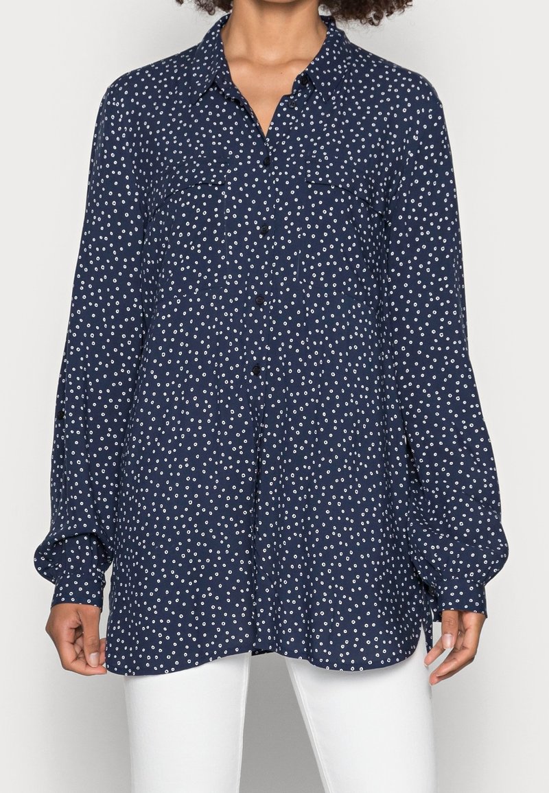 Femme portant une chemise bleu marine à manches longues boutonnée avec de petits motifs circulaires blancs et un pantalon blanc, debout devant un fond uni.