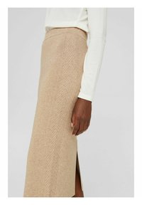 Jupe crayon beige en tricot avec un subtil motif en chevrons, portée avec un haut blanc à manches longues, fente latérale visible près du genou.