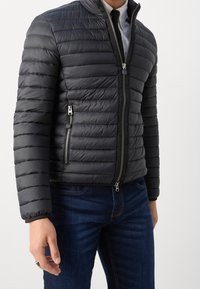 Sort pufferjakke med lynlåslukning foran, med horisontal quiltning, sidelommer og stående krave, kombineret med blå jeans.