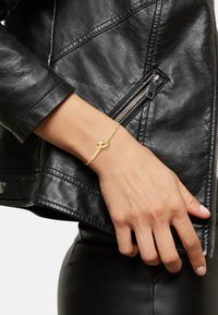 Muñeca con pulsera de nudo dorado llevada sobre la manga de una chaqueta de cuero negro, mano descansando cerca de la cadera con pantalones de cuero negro visibles.