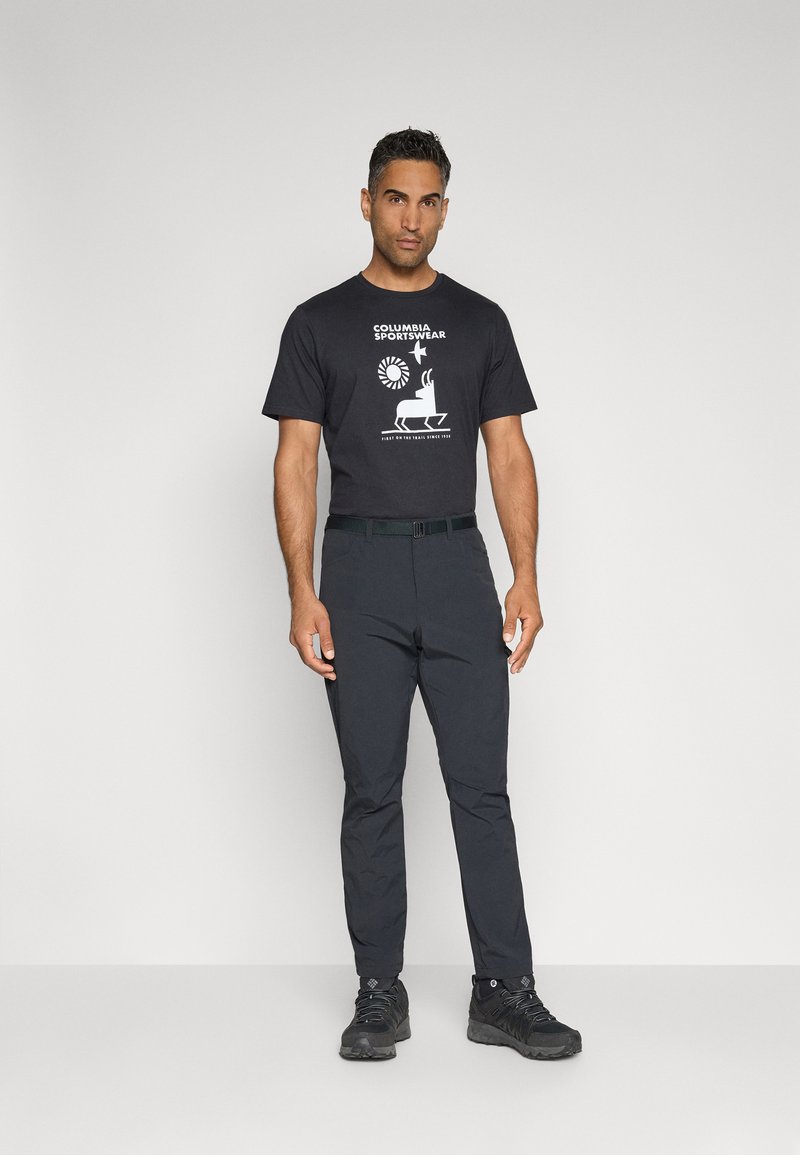 Schwarzes T-Shirt mit kurzen Ärmeln und weißem Logo, kombiniert mit dunkelgrauen Hosen und schwarzen Wanderschuhen, vor einem hellen Hintergrund stehend.