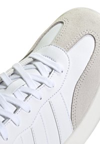 Zapatilla blanca con cuero liso y detalles en ante, con cordones blancos, franjas laterales texturizadas y puntera redondeada.