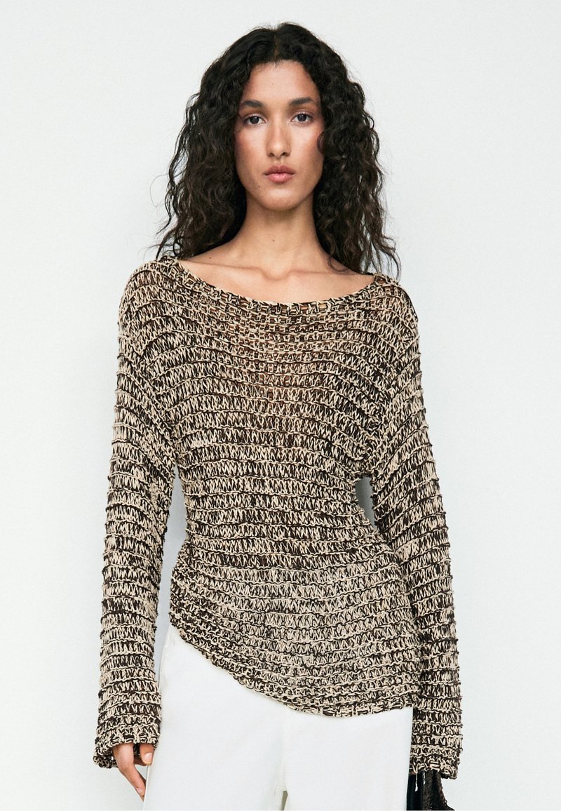 Donna con capelli ricci che indossa un maglione a maglia marrone e beige, morbido e strutturato, e pantaloni bianchi, in piedi davanti a uno sfondo semplice.