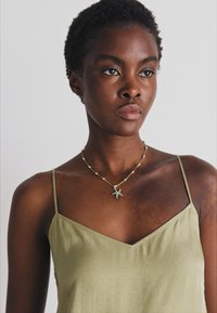 Gul stjerneformet vedhængsnekke på en model. Modellen bærer en lys olivengrøn, ærmeløs top med V-hals.