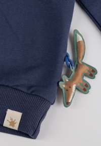 Marineblauer Sweatshirt mit gerippten Bündchen, versehen mit einem Stoff-Fuchs-Label, das mit einem blauen Kordelband befestigt ist. Das Etikett ist mit braunen, weißen und grünen Akzenten gestaltet.