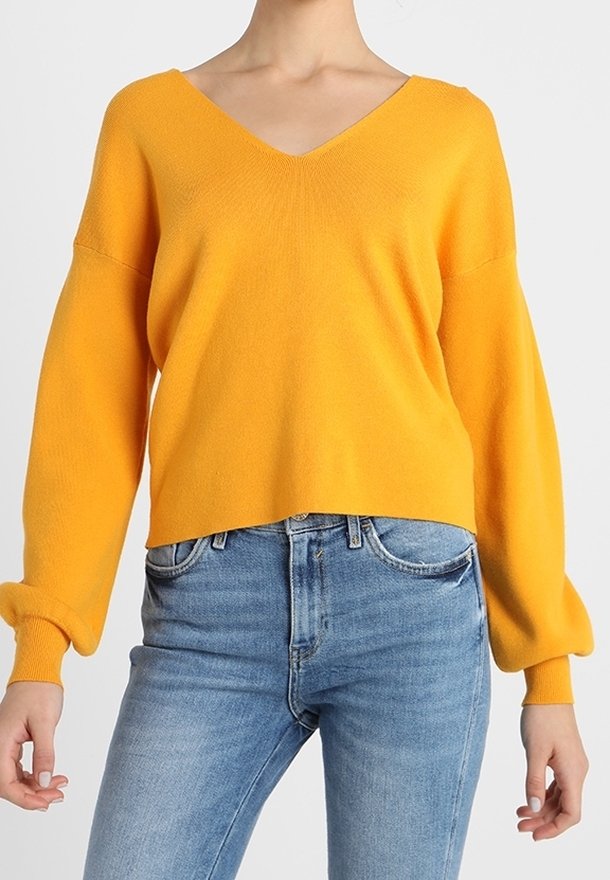 Femme portant un pull jaune vif à col en V avec manches longues, associé à un jean taille haute bleu clair.