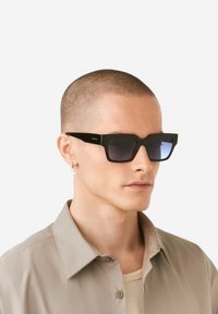 Fekete négyzet alakú napszemüveg, amely gradient sötét lencsékkel rendelkezik, és figyelemfelkeltő keret designnal bír. A karok textúrázottak, és egy minimális márkacímke is látható.