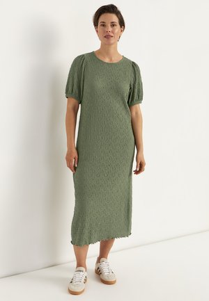 Frau steht vor weißer Wand und trägt ein strukturiertes, olivgrünes Midikleid mit Puffärmeln und weißen Sneakern.