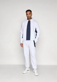 Άνδρας που στέκεται φορώντας λευκή φόρμα με ναυτικές πλευρές, ναυτική μπλούζα και λευκά αθλητικά παπούτσια, σε απλό ανοιχτό γκρίζο φόντο.