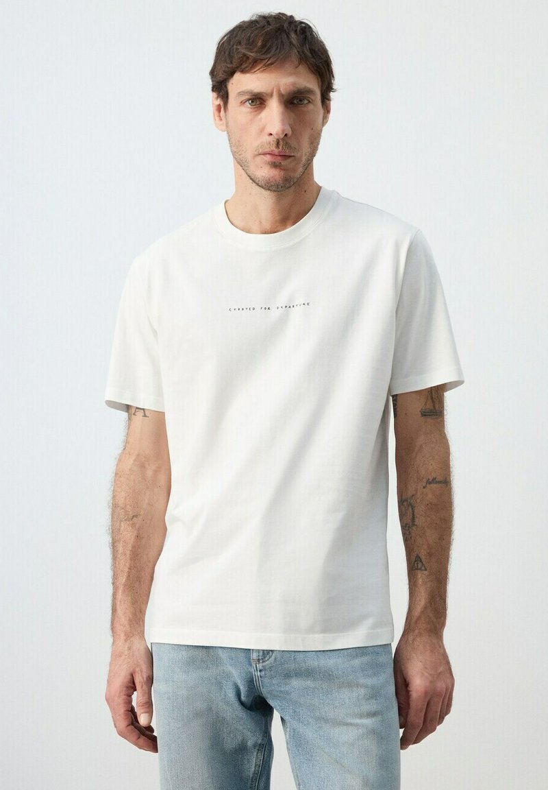 Homme aux cheveux bruns courts portant un T-shirt blanc avec un texte et un jean bleu clair, debout devant un fond clair uni.