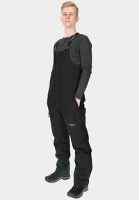 Black waterproof overalls with adjustable straps, multiple zippered pockets, and reinforced seams. Pairing with black footwear, textured fabric.