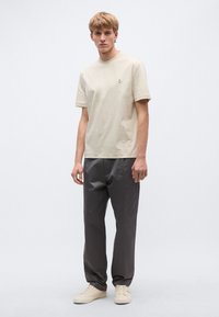 Μπεζ κοντομάνικη μπλούζα με στρογγυλή λαιμόκοψη και μικρό λογότυπο, σε συνδυασμό με σκούρο γκρι παντελόνι και ανοιχτά αθλητικά παπούτσια. Απλός, καθημερινός σχεδιασμός.