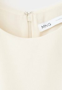 Encolure d’un vêtement couleur crème avec une fermeture éclair visible dans le dos et une étiquette de marque blanche MNG cousue à l’intérieur du col.
