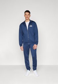 Navy blue tracksuit with zip-up jacket and matching pants, made of smooth fabric. Features ribbed cuffs and a logo on the chest. White sneakers.