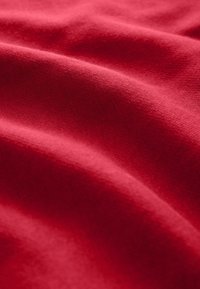 Tessuto di cotone rosso con una consistenza morbida, caratterizzato da onde e pieghe delicate che creano profondità. La superficie presenta un motivo a maglia fine.