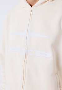 Sudadera beige con cremallera que presenta un logotipo bordado en blanco, capucha ajustable y una textura suave. Primer plano del diseño frontal y la cremallera.