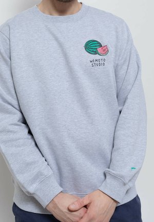 Mężczyzna ubrany w jasnoszarą bluzę z grafiką arbuza i napisem "WEMOTO STUDIO" na klatce piersiowej, z rękami splecionymi z przodu.