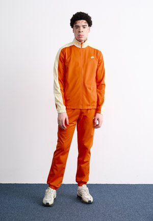 Orange tracksuit with a cream accent on the sleeves, featuring a high collar and elastic cuffs. Paired with light-colored sneakers.