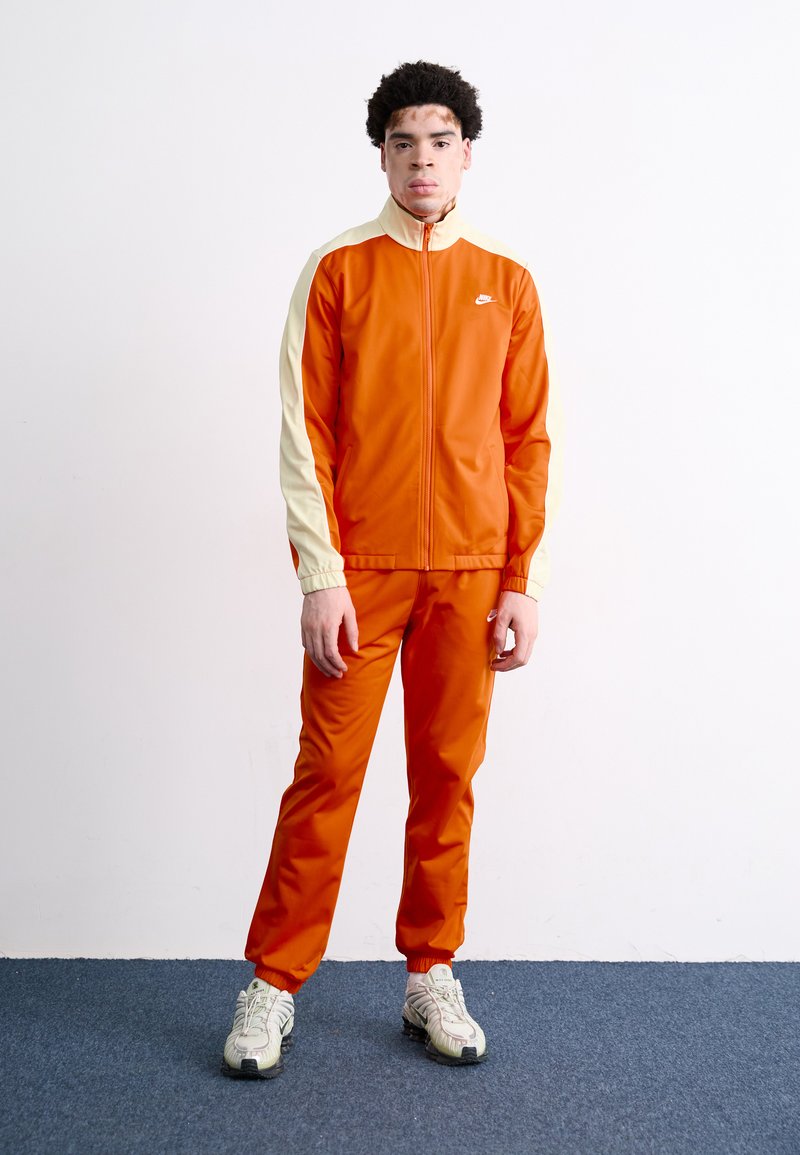 Fato de treino laranja com um detalhe creme nas mangas, apresentando um colarinho alto e punhos elásticos. Combinado com ténis de cor clara.