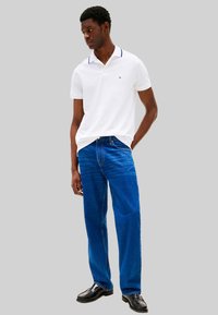 Witte poloshirt met een blauwe kraag en logo, gecombineerd met blauwe jeans met rechte pijpen. Model staat met één hand in de zak en draagt zwarte schoenen.