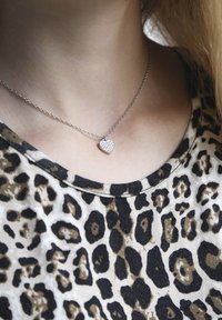 Zilveren halsketting met een klein hartje hangertje versierd met heldere stenen, gedragen boven een shirt met luipaardprint met een ronde hals.