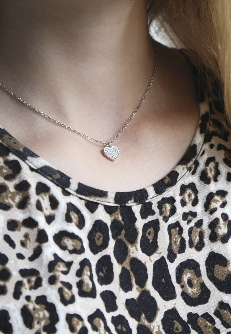 Zilveren halsketting met een klein hartje hangertje versierd met heldere stenen, gedragen boven een shirt met luipaardprint met een ronde hals.